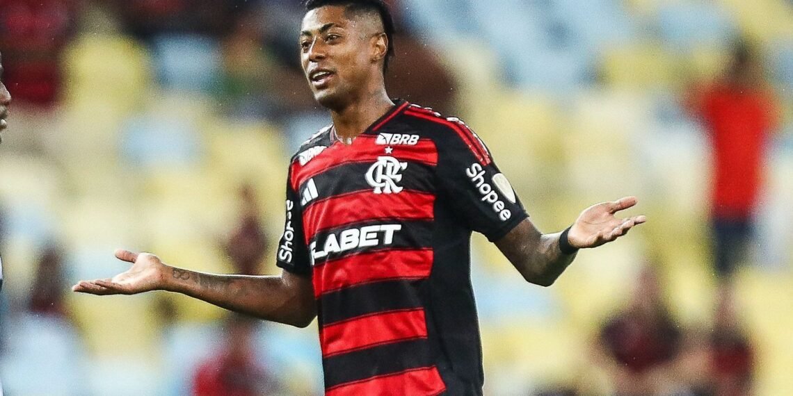 Jogador de futebol em campo, vestindo uniforme do Flamengo, gesticula com os braços abertos, expressão de questionamento.