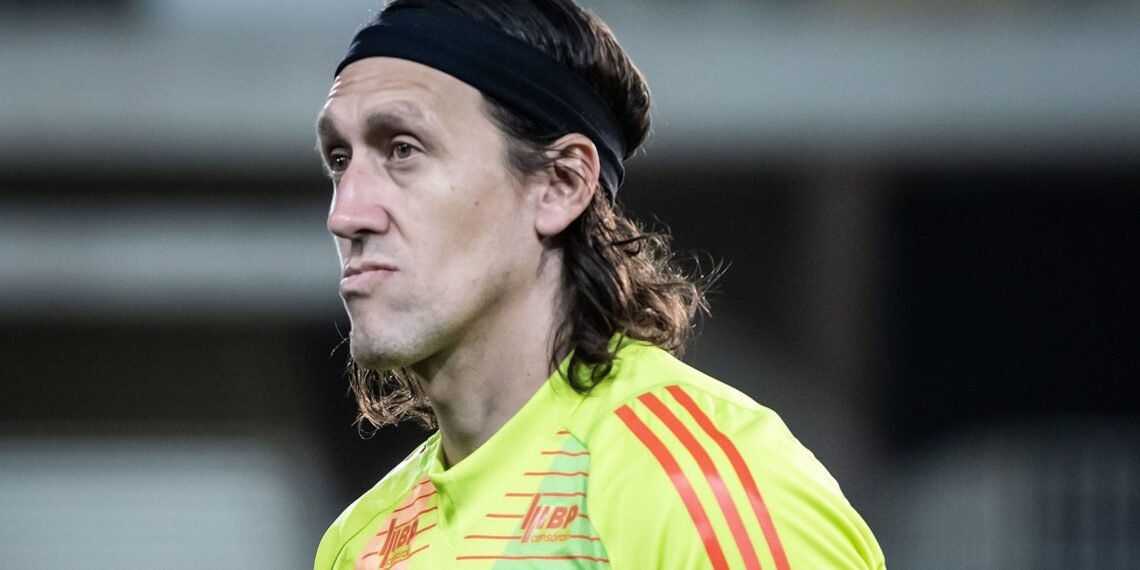 Goleiro de cabelo longo e faixinha preta, usando uniforme amarelo no campo de futebol, expressa concentração.