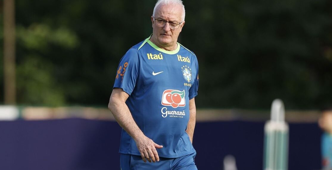 Treinador de futebol em uniforme azul observa treino em campo verdejante, com logotipos de patrocinadores no uniforme.