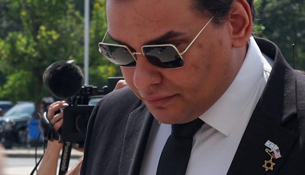 Pessoa com óculos escuros em traje formal caminhando ao ar livre, com microfones ao fundo, sugerindo presença da mídia.