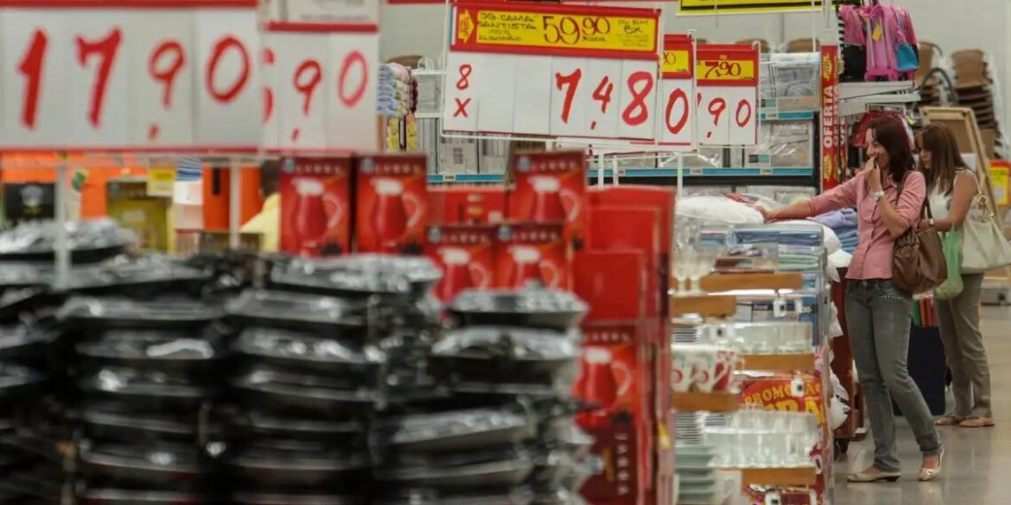 Clientes percorrem corredor de loja, observando promoções em diversas mercadorias, como utensílios domésticos e eletrodomésticos.