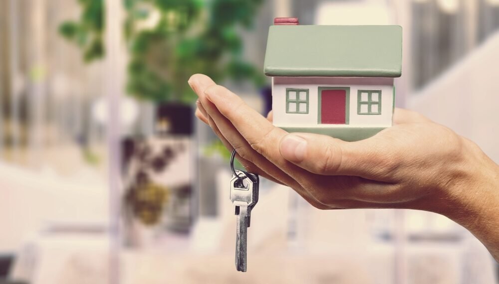 Mão segurando uma miniatura de casa com chave pendurada, simbolizando compra de imóvel ou investimento imobiliário.
