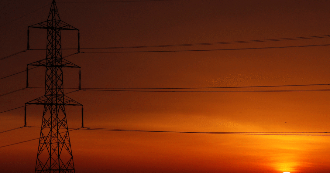 Pôr do sol vibrante em tons de laranja com torre de transmissão de energia ao fundo destacando contrastes urbanos e naturais.