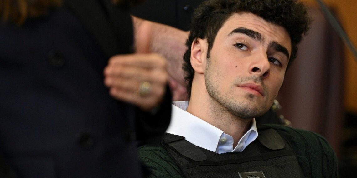 Homem jovem em cena de tribunal, com expressão de interesse, usa colete protetor e está rodeado por outras pessoas.