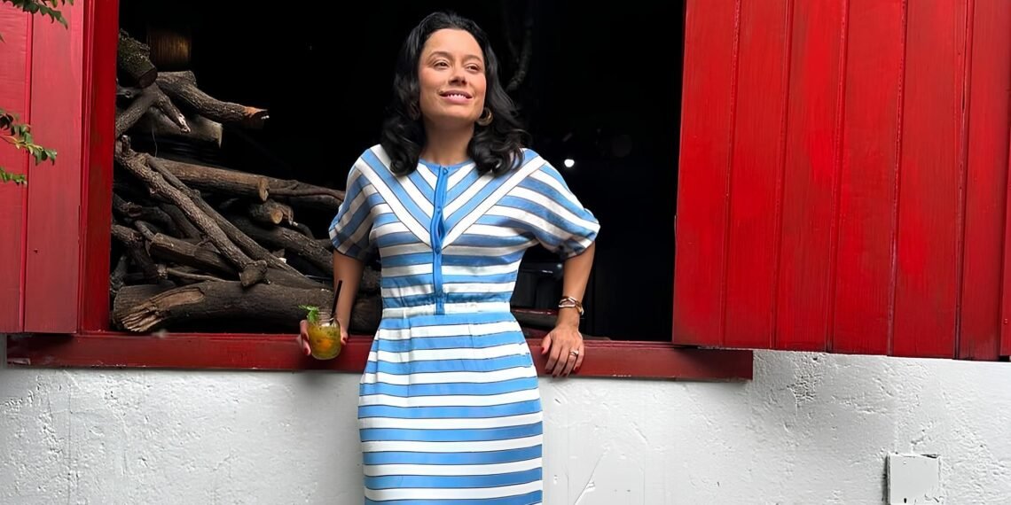 Mulher em vestido listrado azul e branco sorri em frente a pilha de lenha, segurando bebida; cenário rústico com janela vermelha.