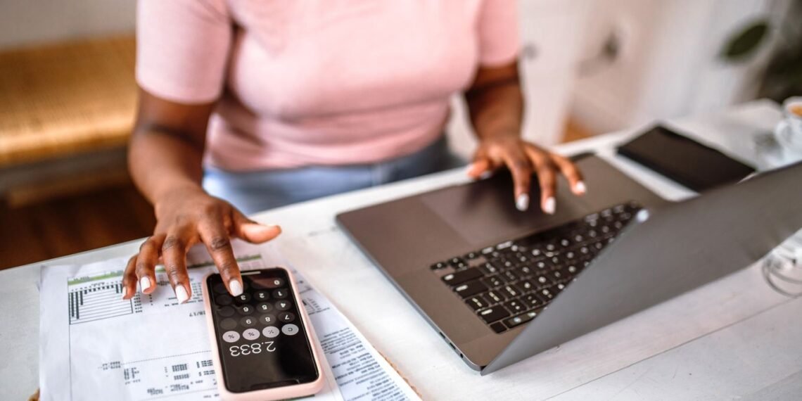 Pessoa em casa usando laptop e calculadora para gerenciar finanças, com planilhas sobre a mesa para controle de despesas.