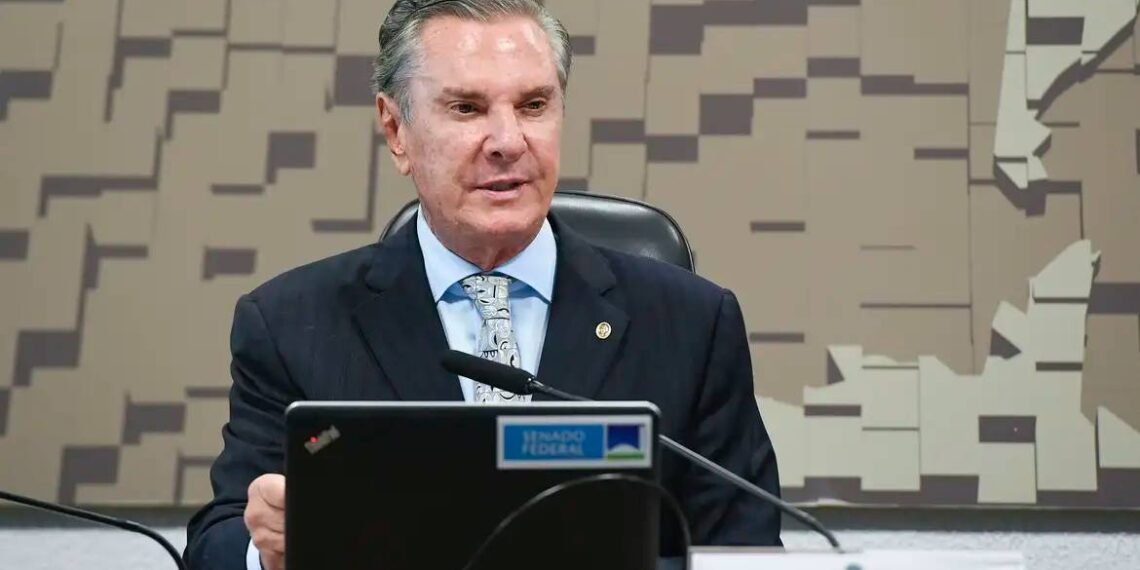 Homem de terno fala em microfone em reunião governamental, com placa do Senado e decoração geométrica ao fundo.