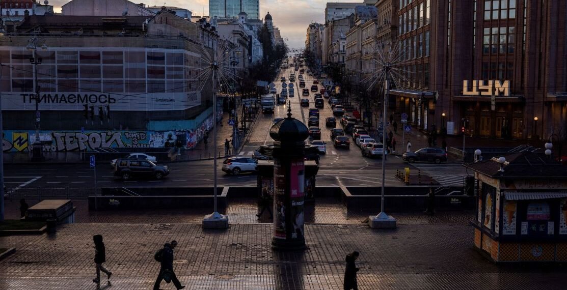 Rua movimentada em centro urbano ao entardecer, com pedestres e carros entre edifícios históricos e modernos.