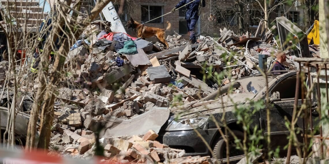 Equipe de resgate busca sobreviventes em destroços, com ajuda de cão farejador, após colapso de prédio.