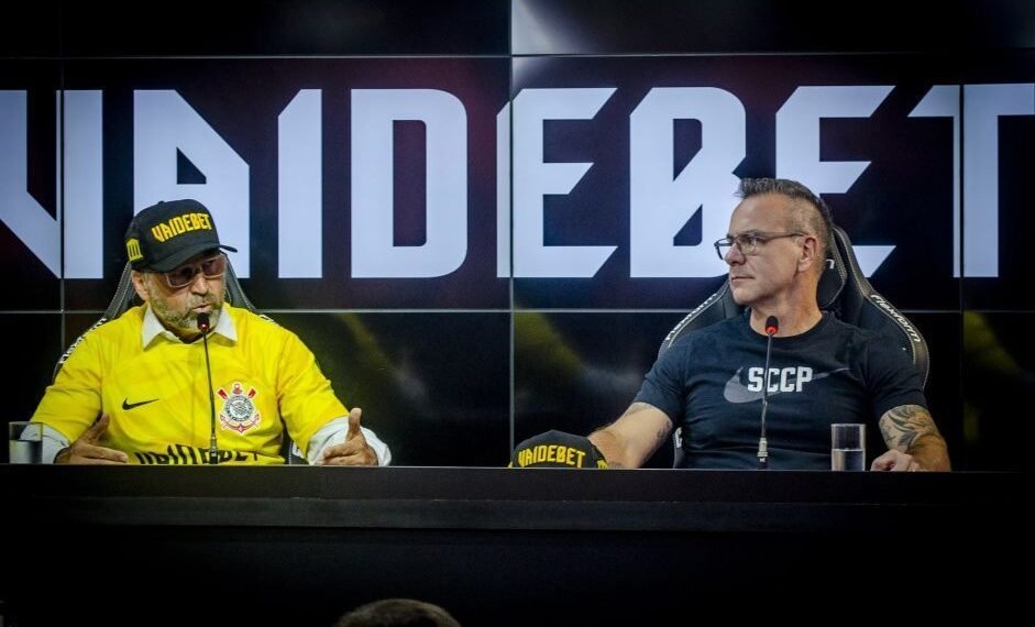 Dois homens em entrevista coletiva no Corinthians, usando boné e camisa do clube, com fundo de painel escrito VAIDEBET.