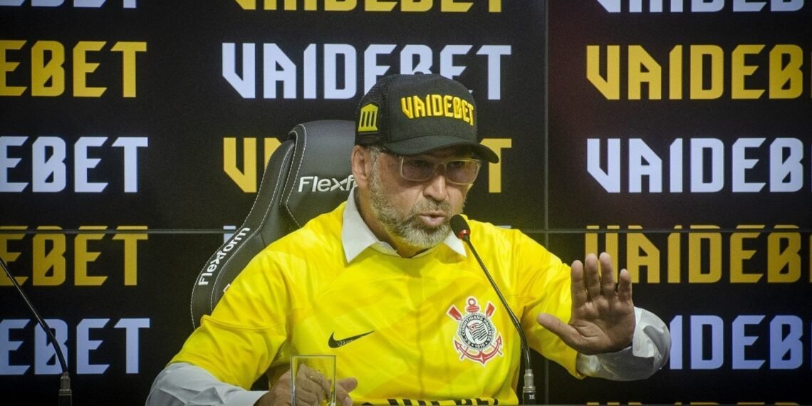 Homem com boné e camisa do Corinthians, em frente a um painel de imprensa VAIDEBET, fala em microfone.
