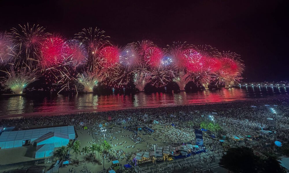 A imagem mostra uma grande queima de fogos de artifício iluminando o céu noturno, com explosões de cores vermelhas e brancas. Abaixo, uma multidão se reúne na praia, com várias pessoas visíveis, e o reflexo dos fogos na água. Ao fundo, há uma estrutura de ponte ou calçadão iluminado.