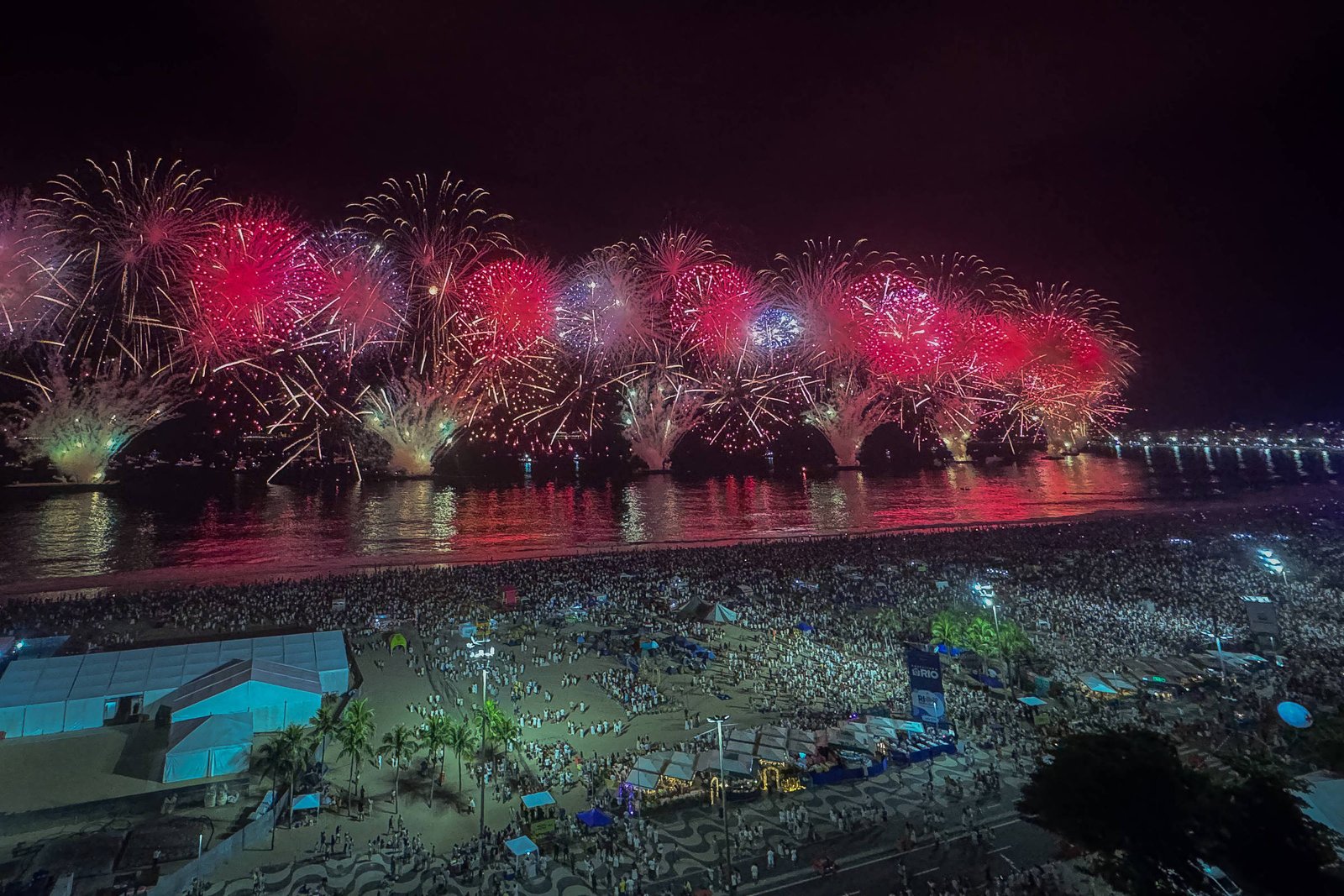 A imagem mostra uma grande queima de fogos de artifício iluminando o céu noturno, com explosões de cores vermelhas e brancas. Abaixo, uma multidão se reúne na praia, com várias pessoas visíveis, e o reflexo dos fogos na água. Ao fundo, há uma estrutura de ponte ou calçadão iluminado.