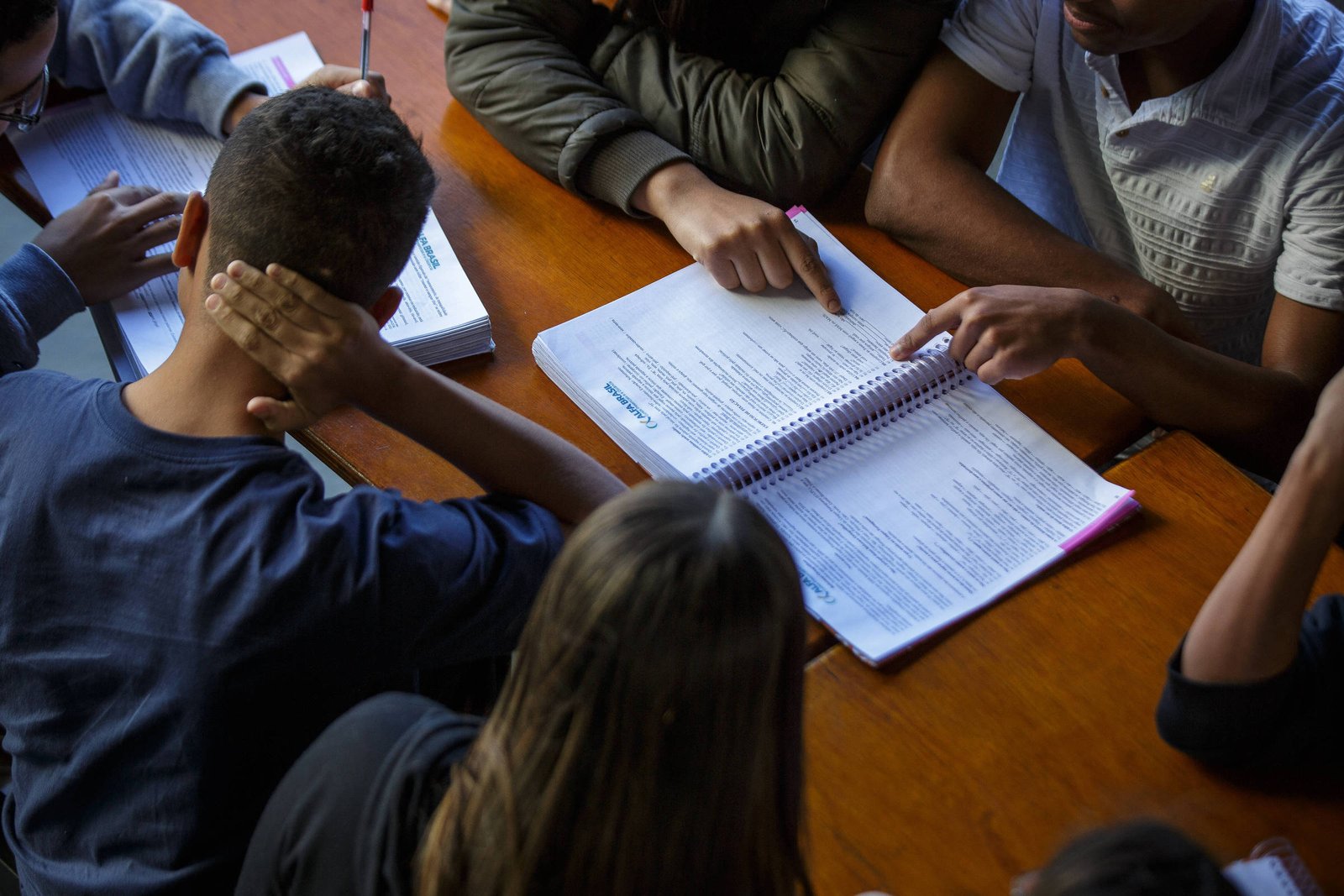 A imagem mostra um grupo de pessoas sentadas ao redor de uma mesa, concentradas em um livro aberto. Algumas mãos estão apontando para o conteúdo do livro, enquanto outras estão apoiadas na mesa. A iluminação é suave, e o ambiente parece ser de estudo ou reunião.