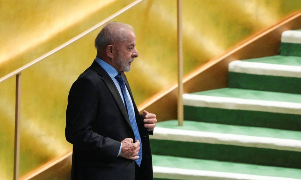 Um homem de terno escuro e gravata azul está em pé em um palco, com uma escada ao fundo. O homem tem cabelo grisalho e uma barba curta. O fundo é dourado e a escada é verde, com degraus cobertos de um material claro. O homem parece estar se preparando para falar ou se apresentar.