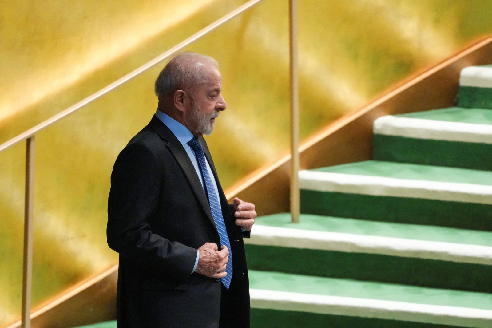 Um homem de terno escuro e gravata azul está em pé em um palco, com uma escada ao fundo. O homem tem cabelo grisalho e uma barba curta. O fundo é dourado e a escada é verde, com degraus cobertos de um material claro. O homem parece estar se preparando para falar ou se apresentar.