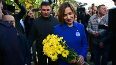 Mulher de casaco azul segura um buquê de flores amarelas enquanto é cercada por várias pessoas e fotógrafos ao ar livre durante o dia.