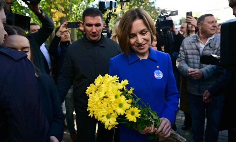 Mulher de casaco azul segura um buquê de flores amarelas enquanto é cercada por várias pessoas e fotógrafos ao ar livre durante o dia.
