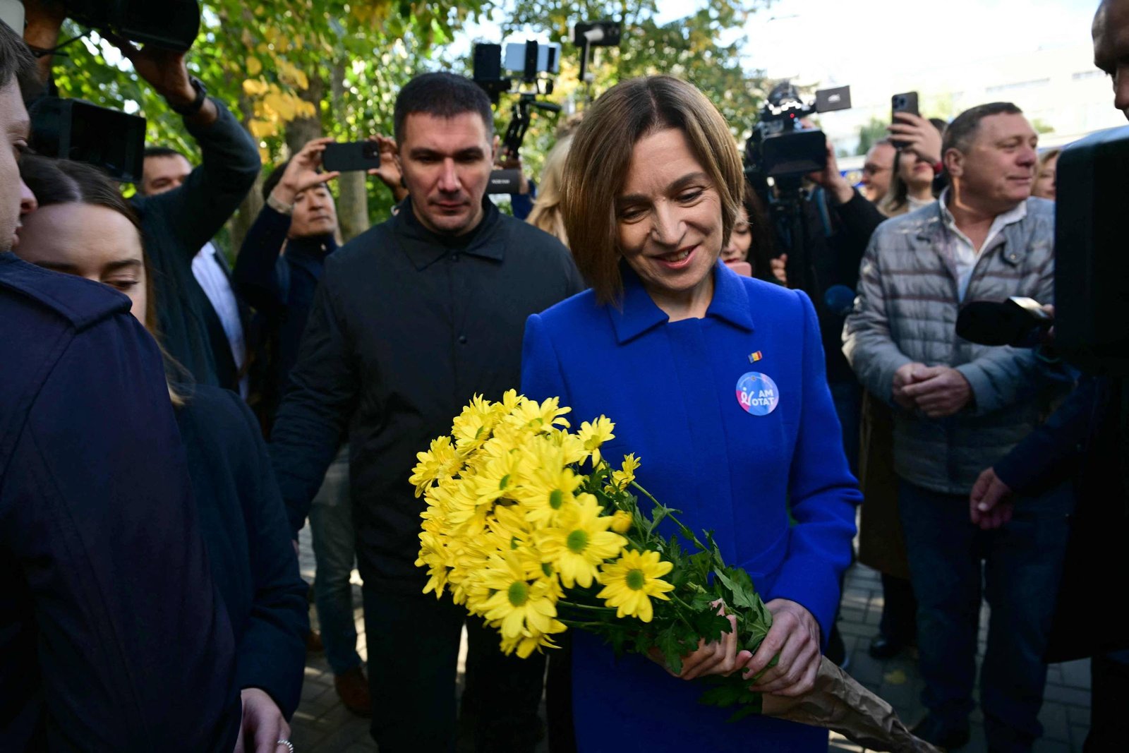 Mulher de casaco azul segura um buquê de flores amarelas enquanto é cercada por várias pessoas e fotógrafos ao ar livre durante o dia.