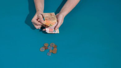 Uma pessoa está segurando notas de dinheiro, com destaque para uma nota de 50, enquanto na mesa há uma pilha de moedas. O fundo é de uma cor azul sólida.