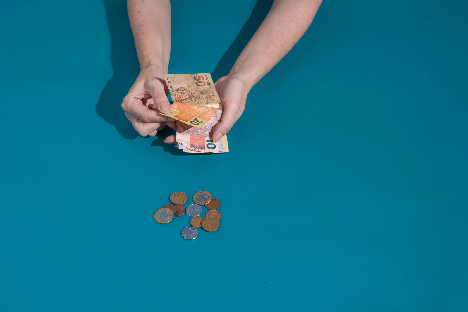 Uma pessoa está segurando notas de dinheiro, com destaque para uma nota de 50, enquanto na mesa há uma pilha de moedas. O fundo é de uma cor azul sólida.