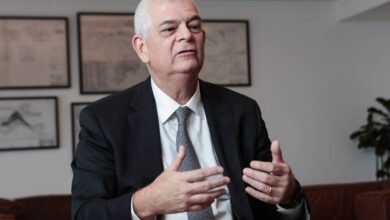 Um homem de terno preto e gravata cinza está sentado em uma mesa, gesticulando com as mãos enquanto fala. Ele tem cabelo grisalho e uma expressão séria. Ao fundo, há quadros com desenhos ou gráficos nas paredes e uma planta em um canto. O ambiente parece ser um escritório moderno.
