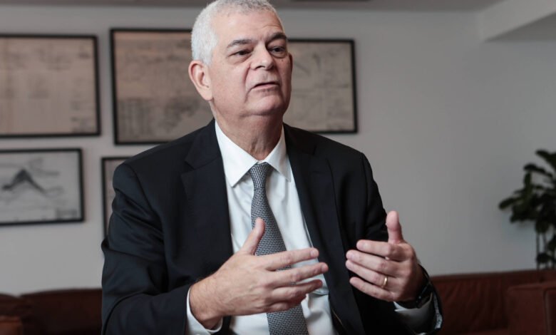 Um homem de terno preto e gravata cinza está sentado em uma mesa, gesticulando com as mãos enquanto fala. Ele tem cabelo grisalho e uma expressão séria. Ao fundo, há quadros com desenhos ou gráficos nas paredes e uma planta em um canto. O ambiente parece ser um escritório moderno.