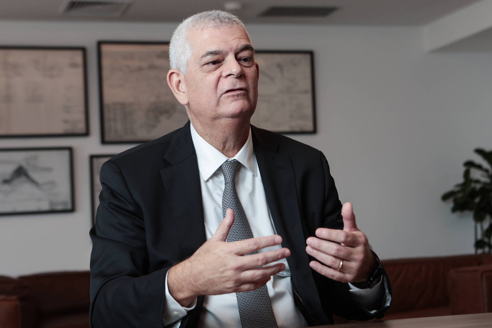 Um homem de terno preto e gravata cinza está sentado em uma mesa, gesticulando com as mãos enquanto fala. Ele tem cabelo grisalho e uma expressão séria. Ao fundo, há quadros com desenhos ou gráficos nas paredes e uma planta em um canto. O ambiente parece ser um escritório moderno.