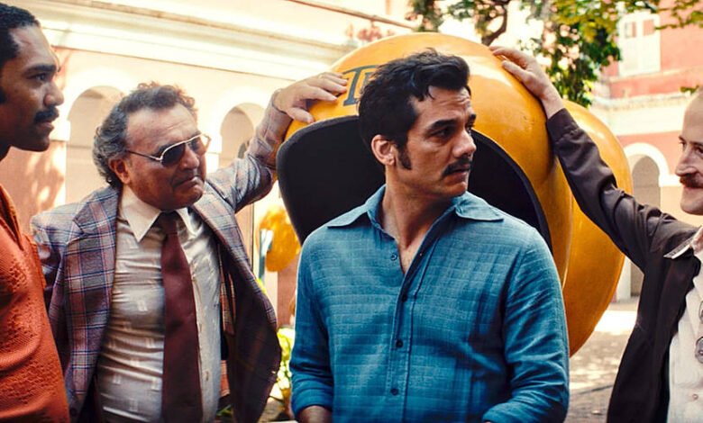 Quatro homens estão em pé em um ambiente externo, possivelmente em um parque ou praça. Eles estão vestidos de maneira casual, com um deles usando uma camisa azul clara e outro uma camisa marrom. O homem no centro parece estar sendo tocado na cabeça por um dos outros. Ao fundo, há uma estrutura grande e arredondada, que parece ser uma escultura ou um objeto decorativo. A iluminação é natural, sugerindo que é um dia ensolarado.