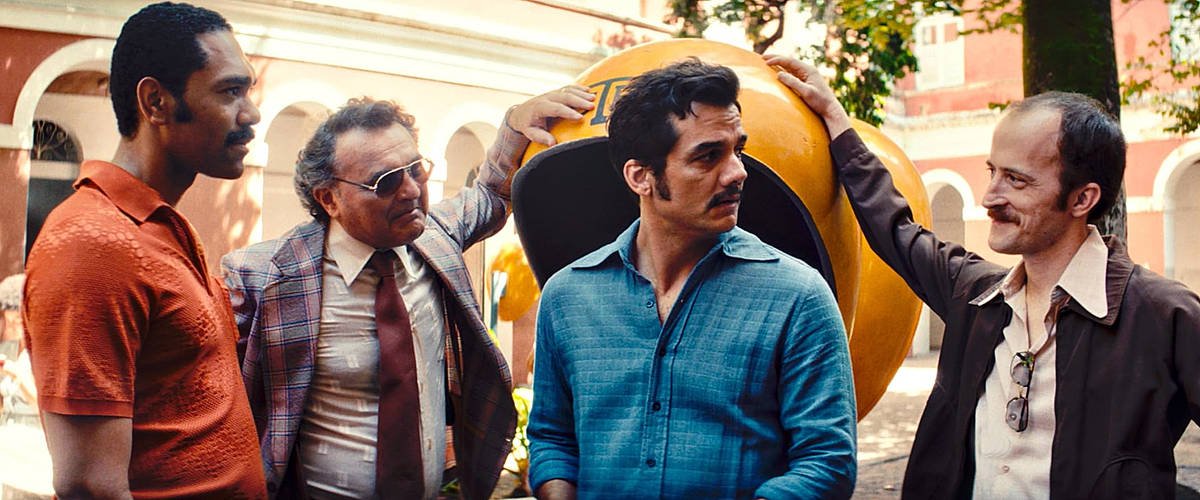 Quatro homens estão em pé em um ambiente externo, possivelmente em um parque ou praça. Eles estão vestidos de maneira casual, com um deles usando uma camisa azul clara e outro uma camisa marrom. O homem no centro parece estar sendo tocado na cabeça por um dos outros. Ao fundo, há uma estrutura grande e arredondada, que parece ser uma escultura ou um objeto decorativo. A iluminação é natural, sugerindo que é um dia ensolarado.