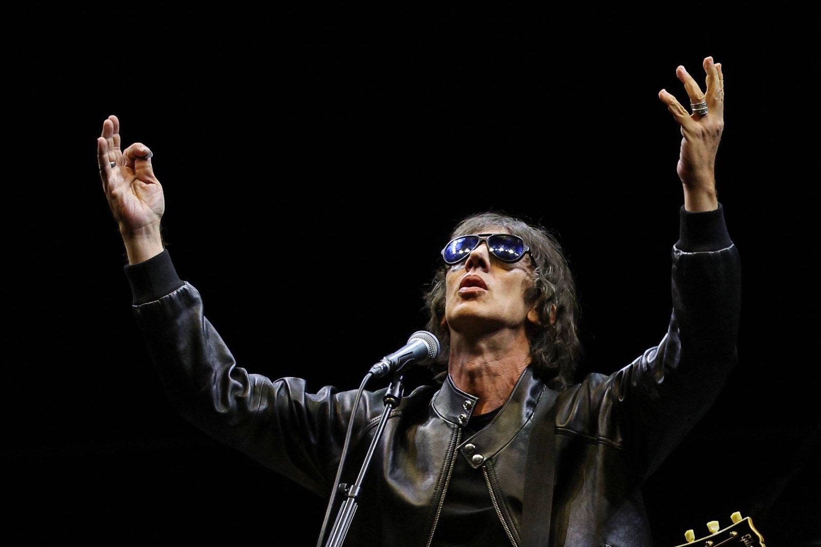 Um artista está se apresentando em um palco, usando óculos escuros e uma jaqueta preta. Ele está com os braços levantados, em uma pose expressiva, enquanto canta em um microfone. Ao fundo, há um cenário escuro, destacando a figura do artista e um violão visível ao lado.