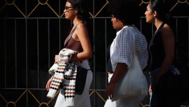 Três mulheres caminham lado a lado em uma rua. A primeira mulher usa óculos e uma blusa de alças, segurando um casaco. A segunda mulher tem cabelo cacheado e usa uma blusa branca com detalhes. A terceira mulher usa uma blusa preta e calças jeans. Ao fundo, há uma grade decorativa com detalhes em dourado.