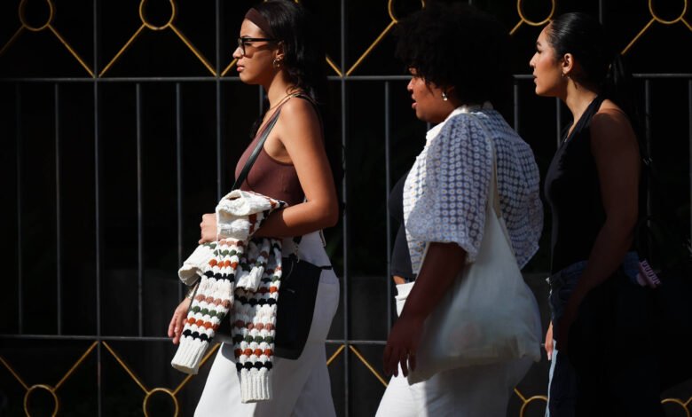 Três mulheres caminham lado a lado em uma rua. A primeira mulher usa óculos e uma blusa de alças, segurando um casaco. A segunda mulher tem cabelo cacheado e usa uma blusa branca com detalhes. A terceira mulher usa uma blusa preta e calças jeans. Ao fundo, há uma grade decorativa com detalhes em dourado.
