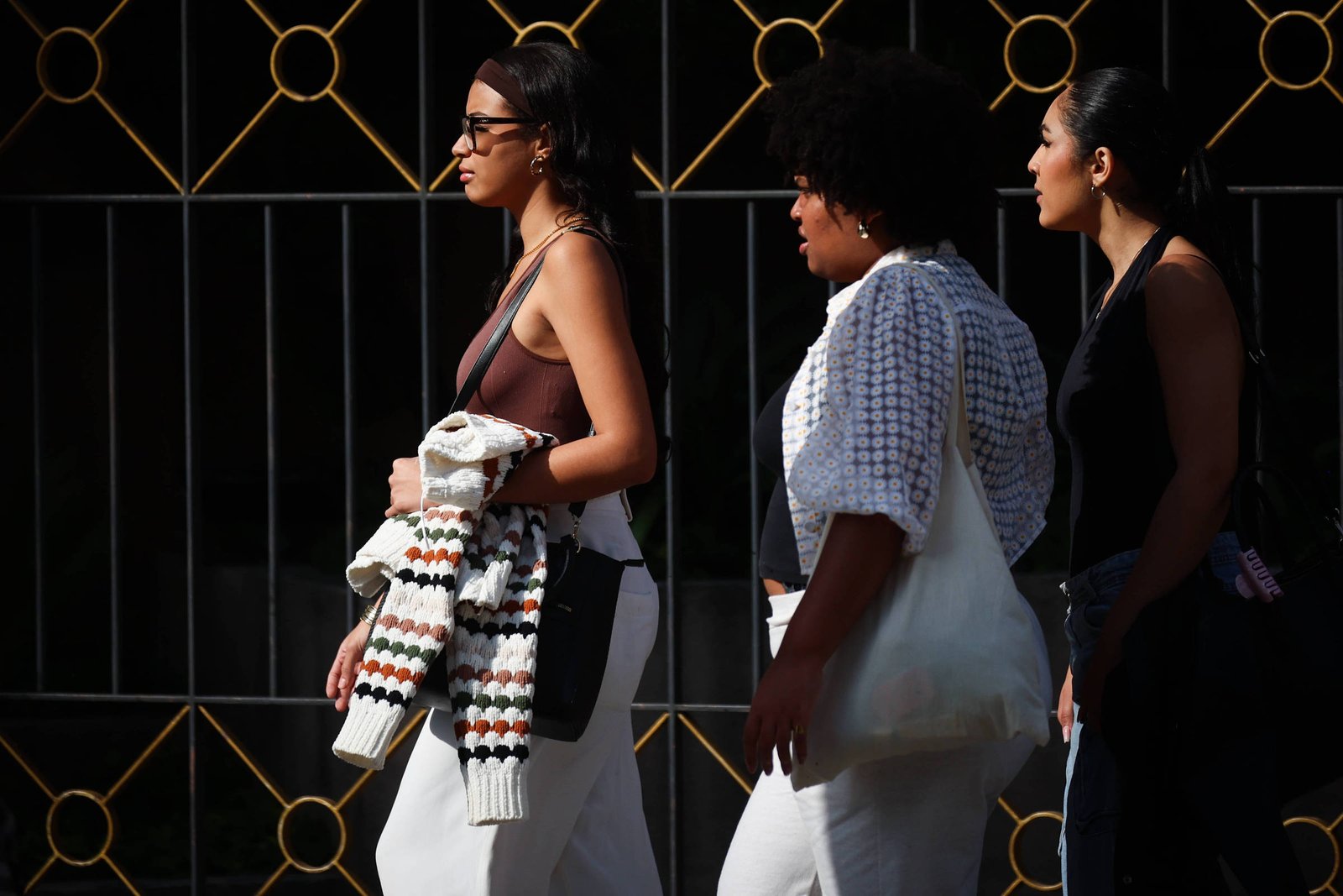 Três mulheres caminham lado a lado em uma rua. A primeira mulher usa óculos e uma blusa de alças, segurando um casaco. A segunda mulher tem cabelo cacheado e usa uma blusa branca com detalhes. A terceira mulher usa uma blusa preta e calças jeans. Ao fundo, há uma grade decorativa com detalhes em dourado.