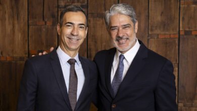 A imagem mostra dois homens sorrindo, posando juntos. O homem à esquerda está usando um terno escuro com uma gravata listrada e uma camisa branca. O homem à direita está vestido com um terno escuro e uma gravata com padrão. Ambos têm cabelos curtos e estão em um ambiente com fundo de madeira. Eles parecem estar em um evento formal.
