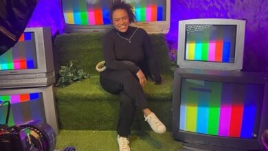 Pessoa sentada em sofá verde cercada por cinco televisores antigos exibindo tela de teste colorida. Ambiente iluminado com luz roxa e decoração com folhas na parede.
