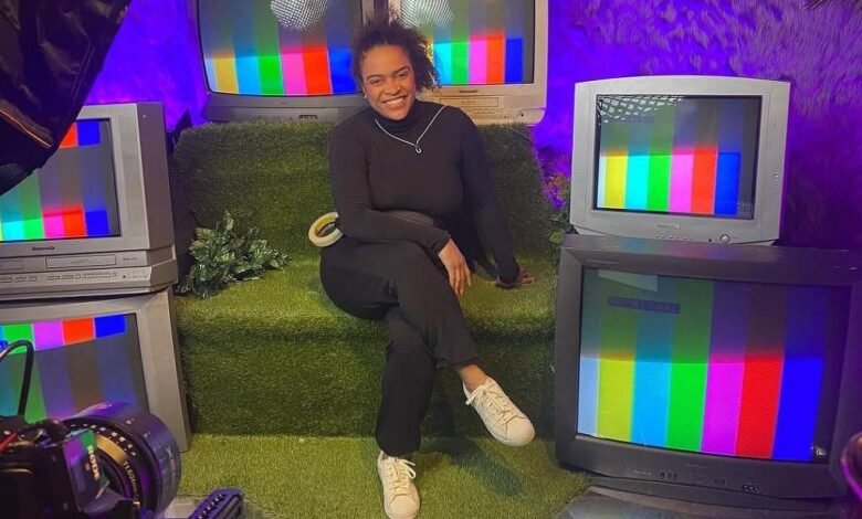 Pessoa sentada em sofá verde cercada por cinco televisores antigos exibindo tela de teste colorida. Ambiente iluminado com luz roxa e decoração com folhas na parede.