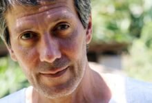 Homem adulto com cabelo curto e grisalho, barba por fazer, olhando diretamente para a câmera. Fundo desfocado com vegetação e luz natural.