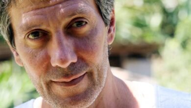 Homem adulto com cabelo curto e grisalho, barba por fazer, olhando diretamente para a câmera. Fundo desfocado com vegetação e luz natural.