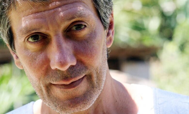 Homem adulto com cabelo curto e grisalho, barba por fazer, olhando diretamente para a câmera. Fundo desfocado com vegetação e luz natural.
