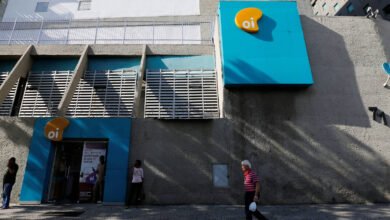Fachada cinza com detalhes azuis da loja Oi em área urbana. Três pessoas estão próximas à entrada e um homem caminha na calçada em frente. Sinalização amarela e branca da Oi aparece na parede.