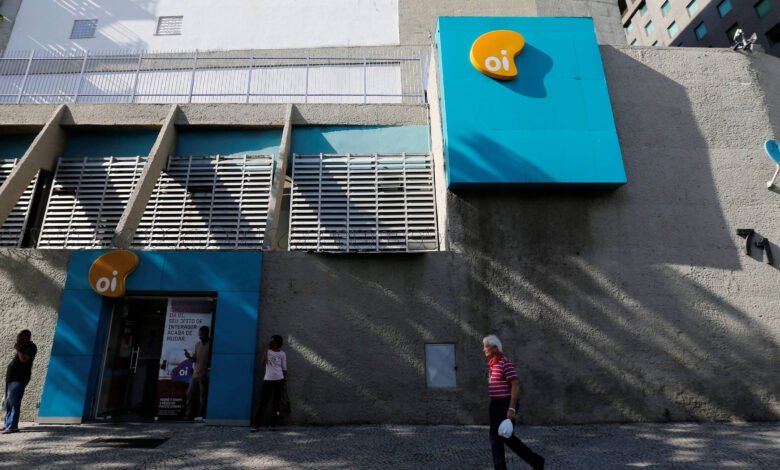 Fachada cinza com detalhes azuis da loja Oi em área urbana. Três pessoas estão próximas à entrada e um homem caminha na calçada em frente. Sinalização amarela e branca da Oi aparece na parede.