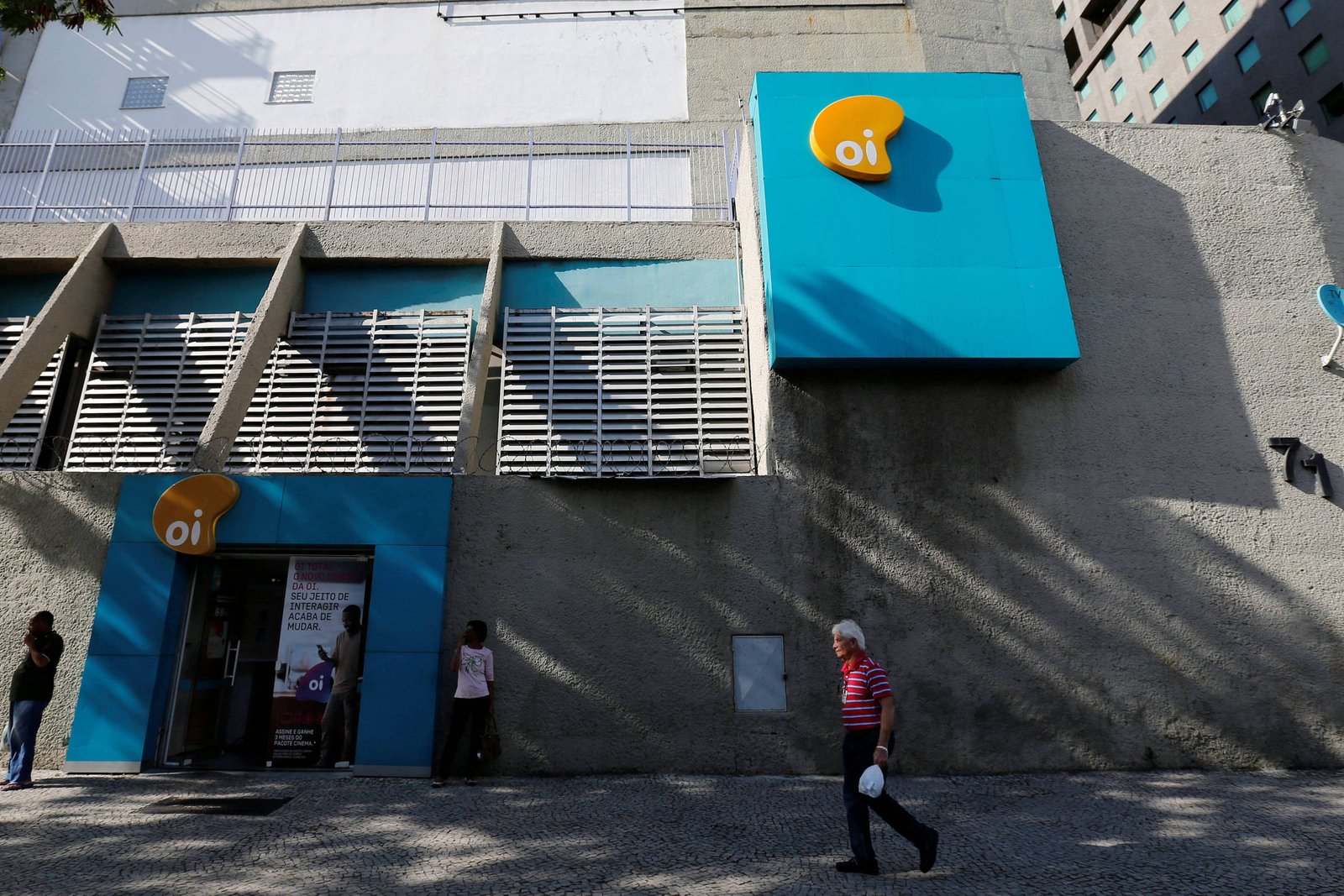 Fachada cinza com detalhes azuis da loja Oi em área urbana. Três pessoas estão próximas à entrada e um homem caminha na calçada em frente. Sinalização amarela e branca da Oi aparece na parede.