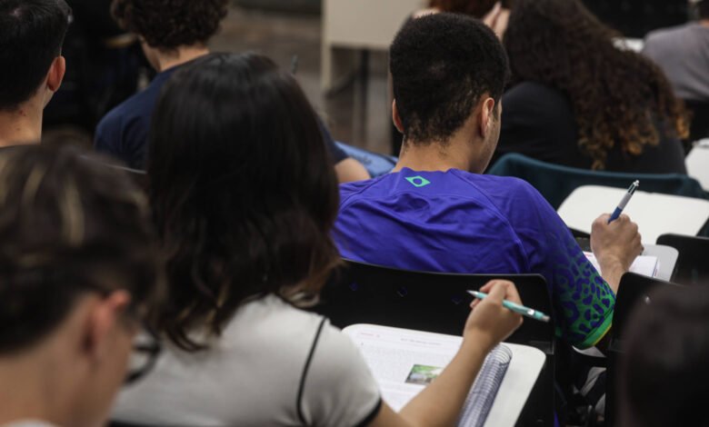 Vários estudantes sentados em carteiras escolares escrevendo em cadernos. Ambiente interno com foco em um jovem de camiseta azul com detalhes verdes.