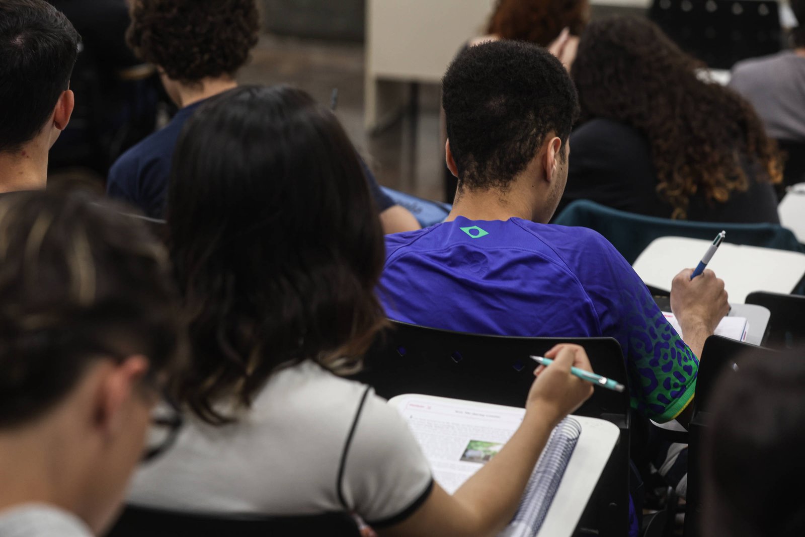 Vários estudantes sentados em carteiras escolares escrevendo em cadernos. Ambiente interno com foco em um jovem de camiseta azul com detalhes verdes.