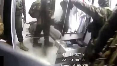 Quatro agentes com roupas camufladas e mochilas estão em um ambiente interno branco. Um deles está com o braço estendido, possivelmente apontando ou segurando algo. Equipamentos e caixas estão visíveis ao redor. Dados de localização e velocidade aparecem sobrepostos na imagem.
