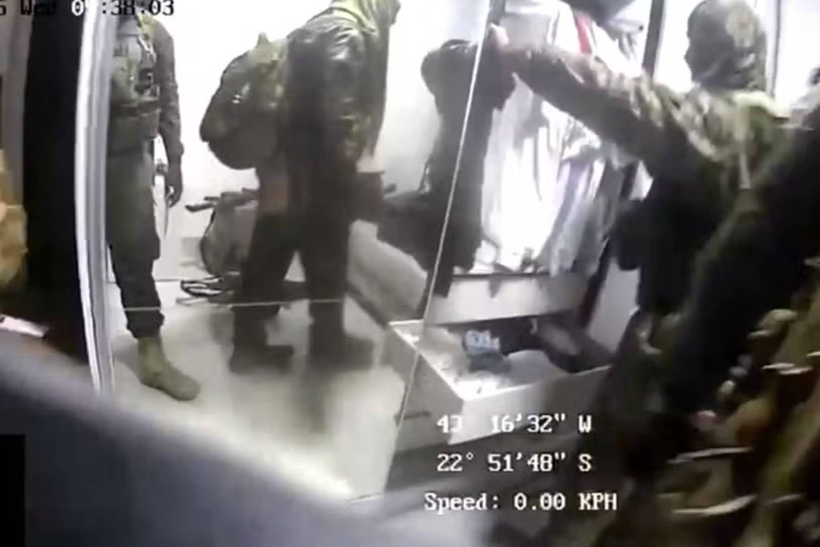 Quatro agentes com roupas camufladas e mochilas estão em um ambiente interno branco. Um deles está com o braço estendido, possivelmente apontando ou segurando algo. Equipamentos e caixas estão visíveis ao redor. Dados de localização e velocidade aparecem sobrepostos na imagem.