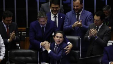 Deputados em trajes formais se reúnem em plenário. Um homem sentado é cumprimentado e abraçado por outro, enquanto colegas ao redor aplaudem.