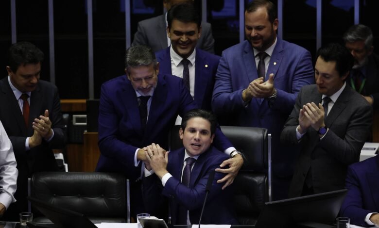 Deputados em trajes formais se reúnem em plenário. Um homem sentado é cumprimentado e abraçado por outro, enquanto colegas ao redor aplaudem.
