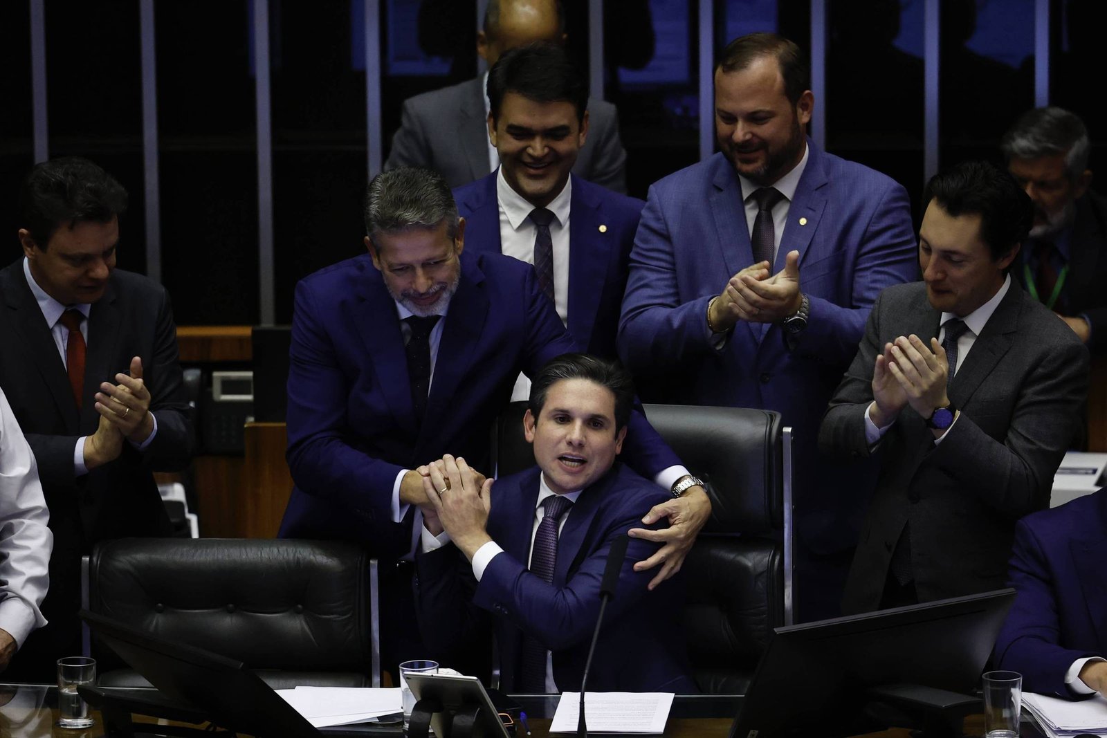 Deputados em trajes formais se reúnem em plenário. Um homem sentado é cumprimentado e abraçado por outro, enquanto colegas ao redor aplaudem.